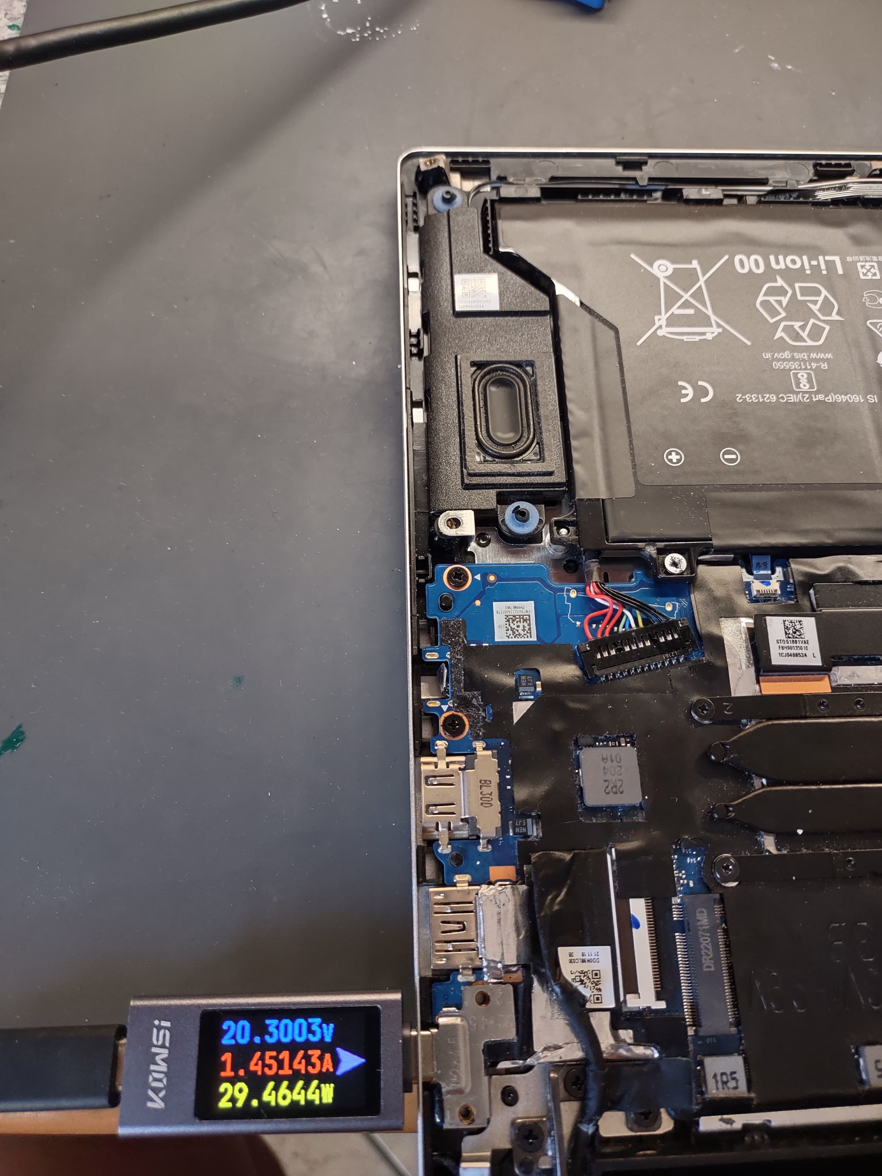 Notebook aperto sul banco del laboratorio in Viale Sarca a Milano dopo la sostituzione del connettore di ricarica in microsaldatura: tester USB-C che segna 20.3V, 1.45A e 29.46W, batteria Li-ion, scheda madre, SSD M.2 e antenne WiFi